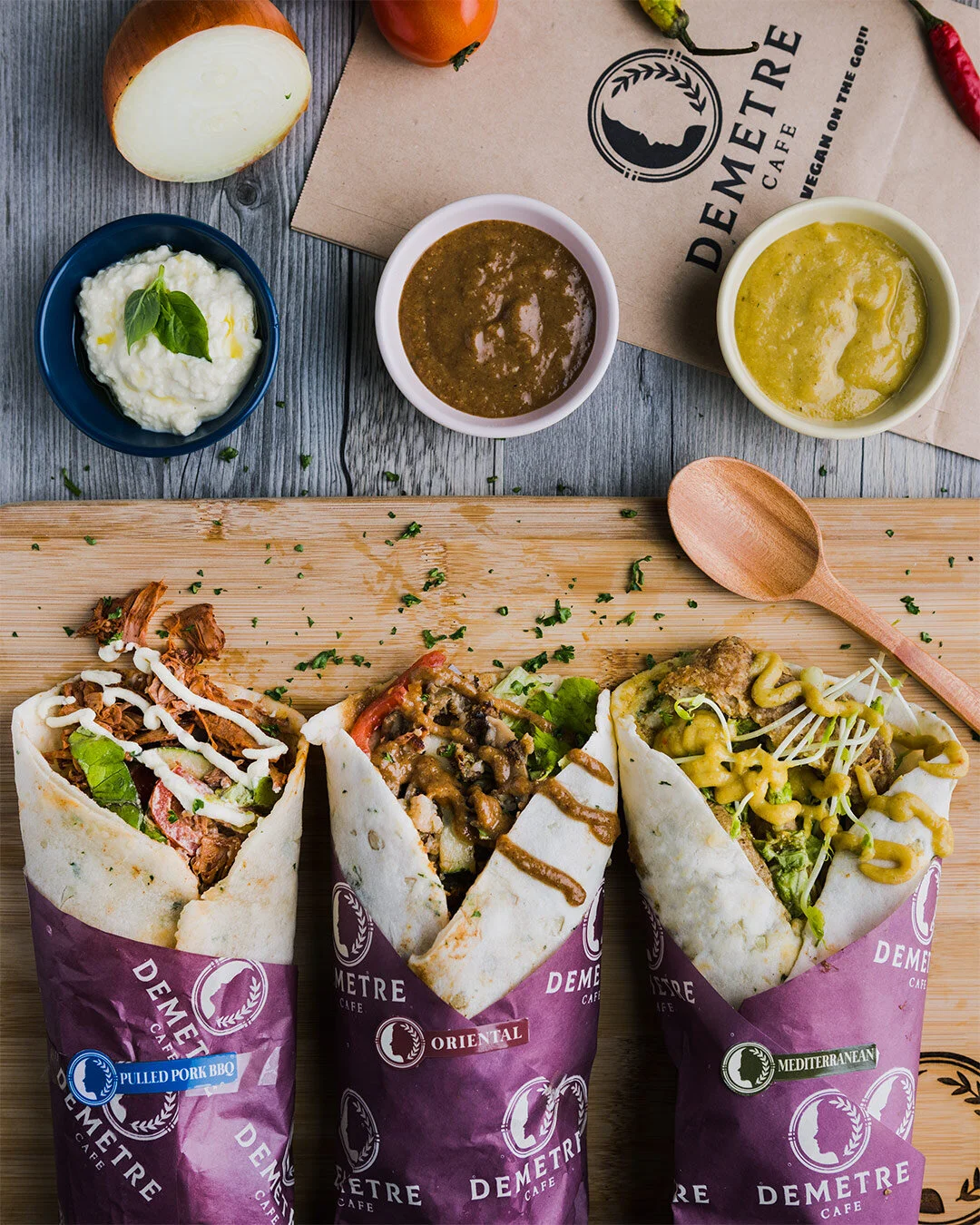 Three burritos on a wooden board with various toppings and sauces in small bowls on a table. There is a partially sliced onion and a paper bag with the Demetre Cafe logo in the background.