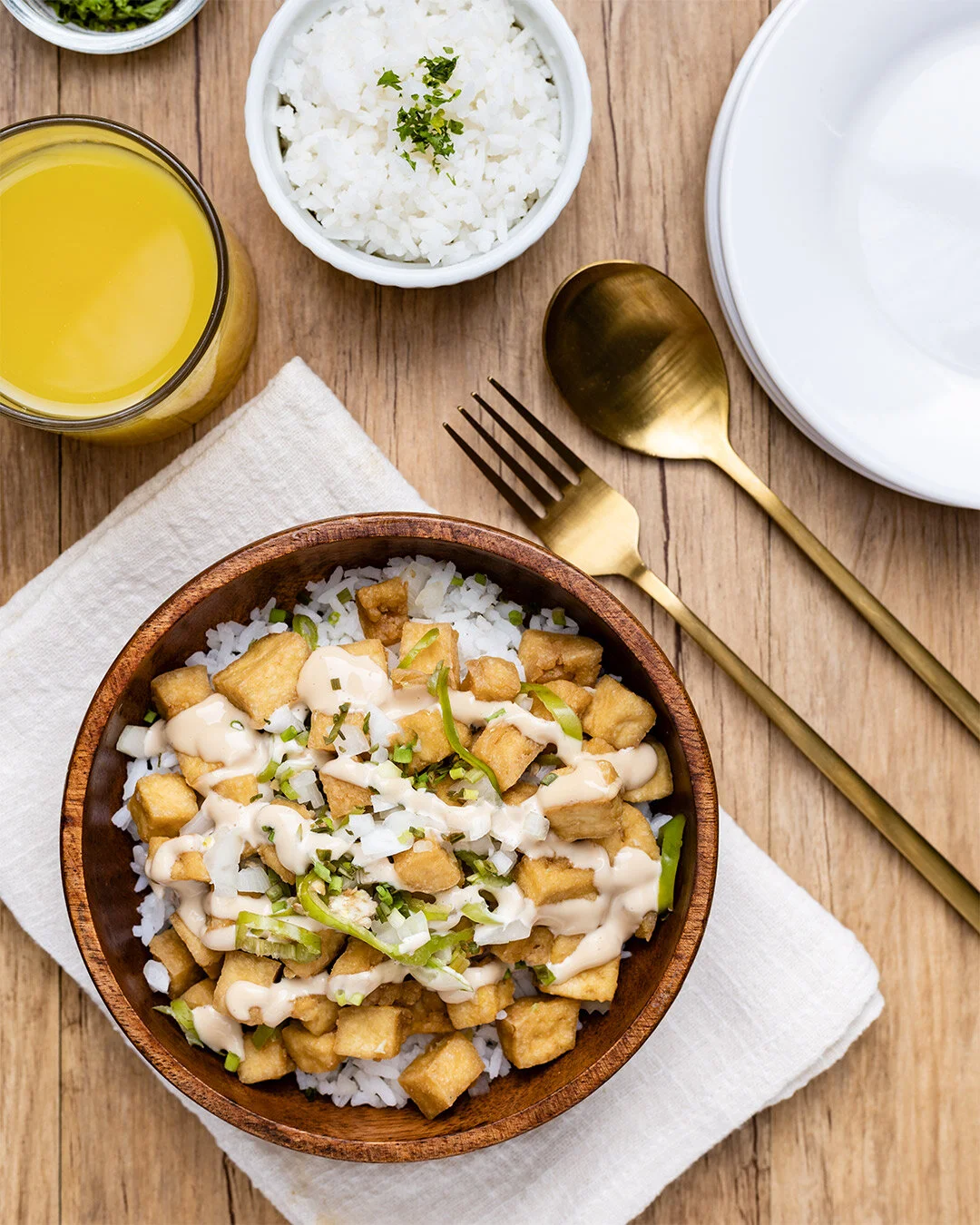 Bowl of rice topped with fried tofu, green onions, and sauce, with side dishes of rice, juice, and salad on a wooden table.