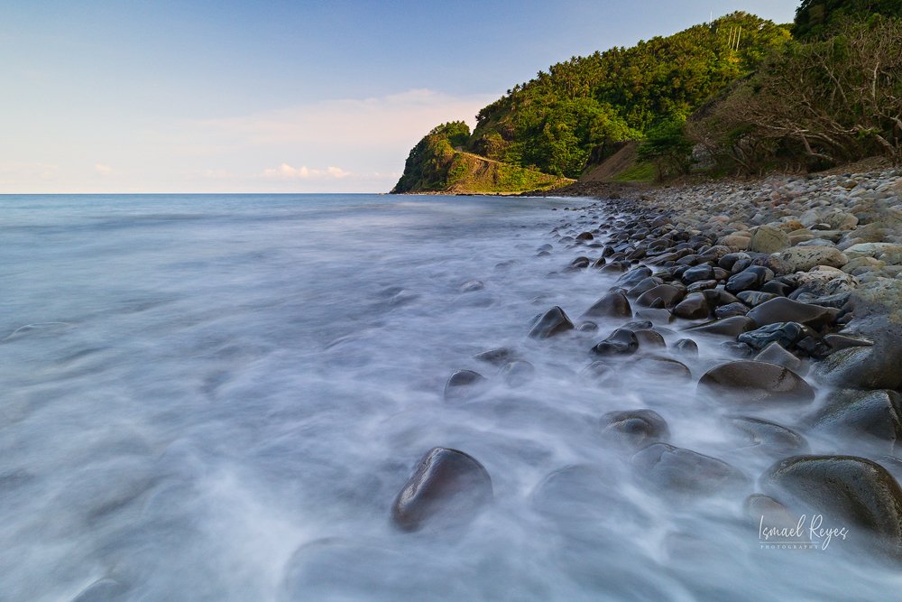 A rocky beach with smooth stones and a forested hill in the background, with ocean waves gently washing over the rocks under a partly cloudy sky.
