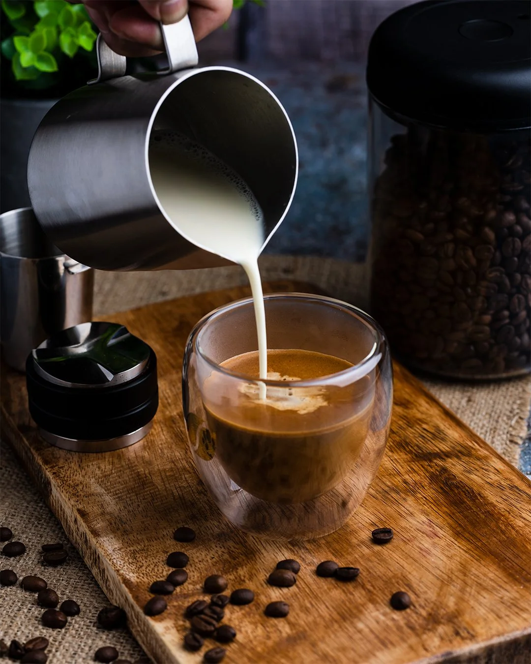Pouring steamed milk into a cup of brewed coffee on a wooden board with coffee beans around, a jar of coffee beans, and a small container nearby.
