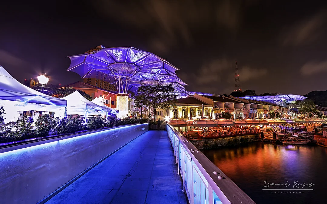 Night view of a lit-up waterfront walkway with colorful buildings, futuristic umbrella-like structures, and boats docked along the water, illuminated by vibrant blue and red lights.