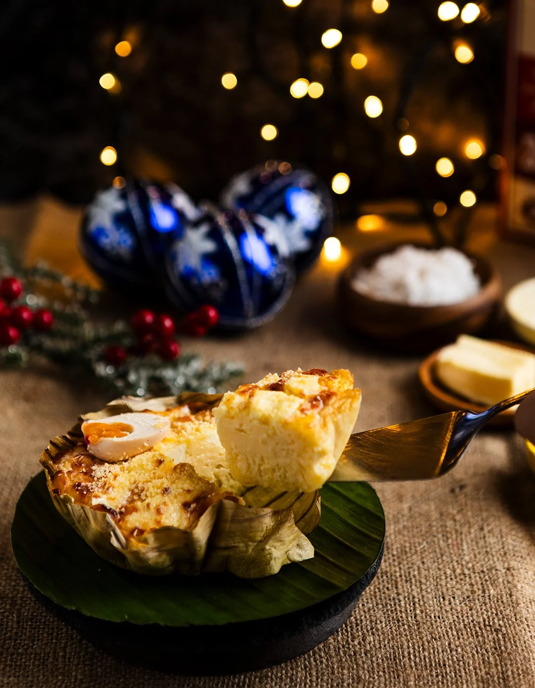 A slice of cheesy baked casserole with a boiled egg on top, served in a cup-shaped baking dish on a black plate, with a festive, Christmas-themed background including a decorated tree with warm lights, Christmas ornaments, berries, and dishes of butt