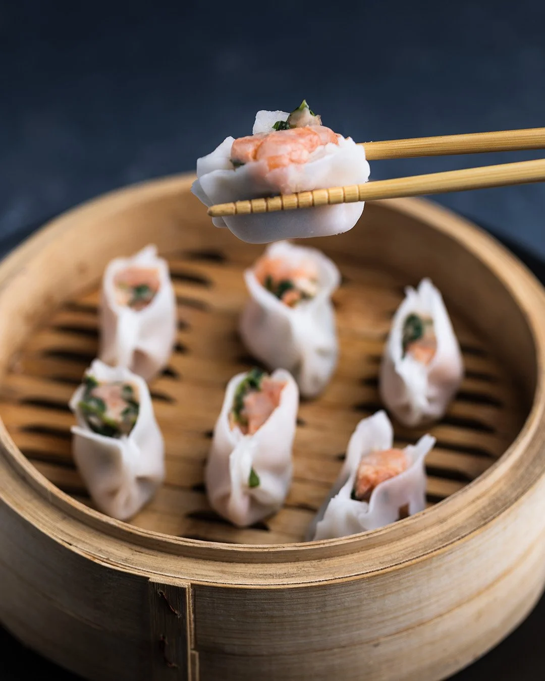 Close-up of a bamboo steamer with shrimp spring rolls and one spring roll being picked up with chopsticks.