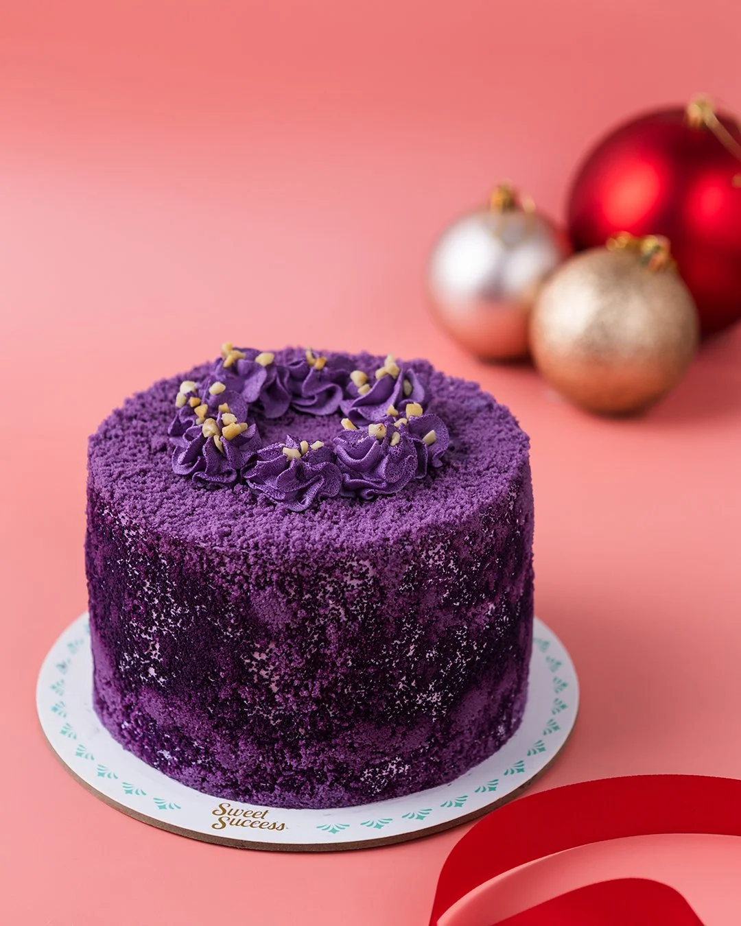 A purple velvet cake decorated with purple frosting and small white sprinkles, placed on a white cake board with a pink background and Christmas ornaments in the background.