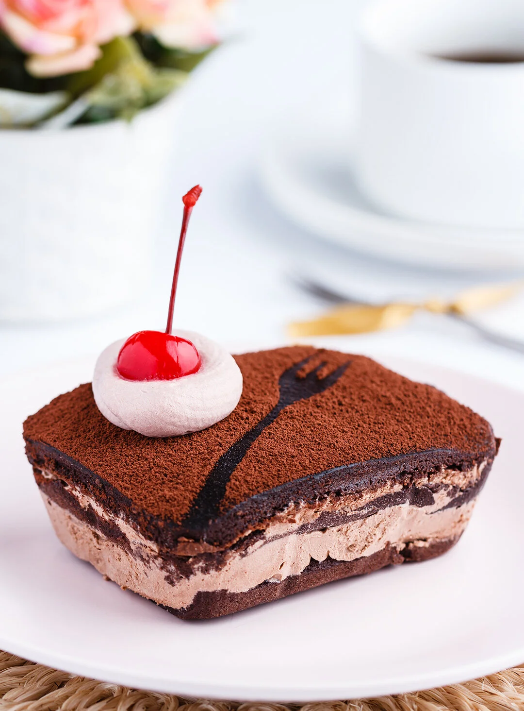 Square slice of tiramisu dessert topped with a maraschino cherry, a dollop of whipped cream, and a chocolate fork design, on a white plate.