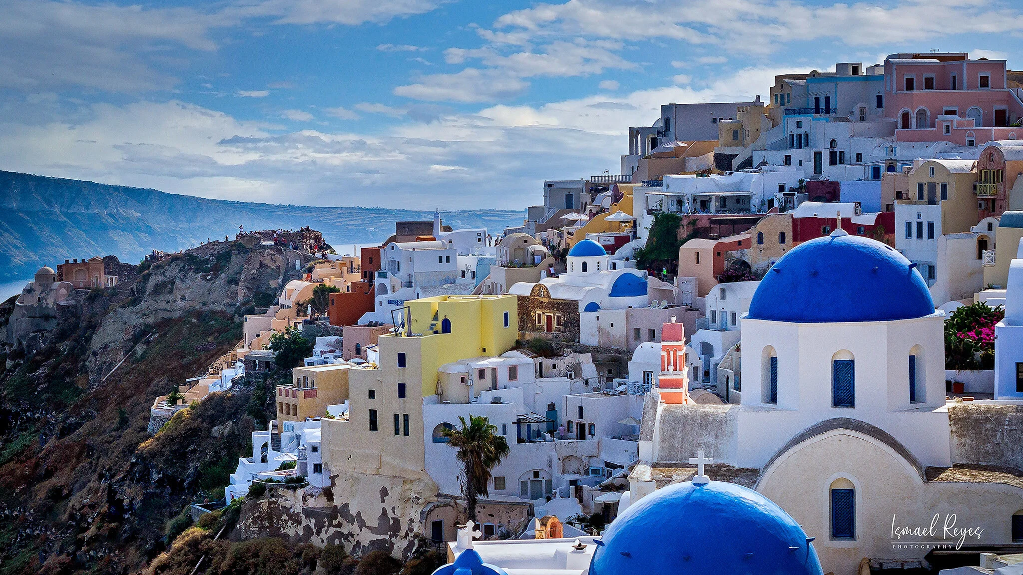 Colorful white, yellow, pink, and blue buildings with domed roofs on a hillside overlooking the sea, in Santorini, Greece.