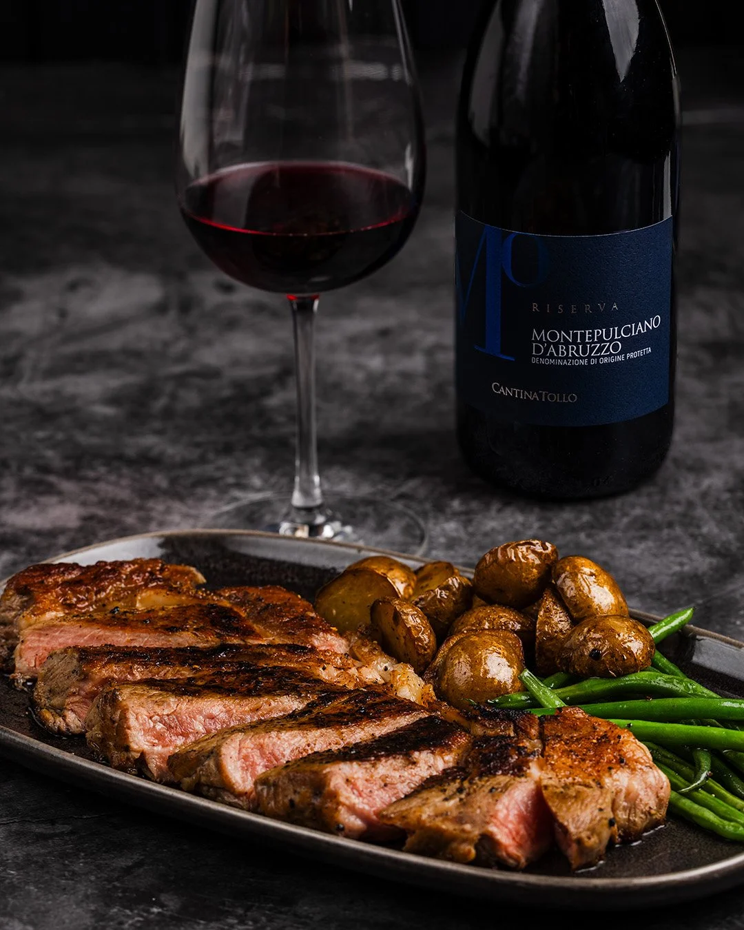 A plate with sliced grilled steak, roasted potatoes, and green beans on a dark surface, with a glass of red wine and a wine bottle in the background.