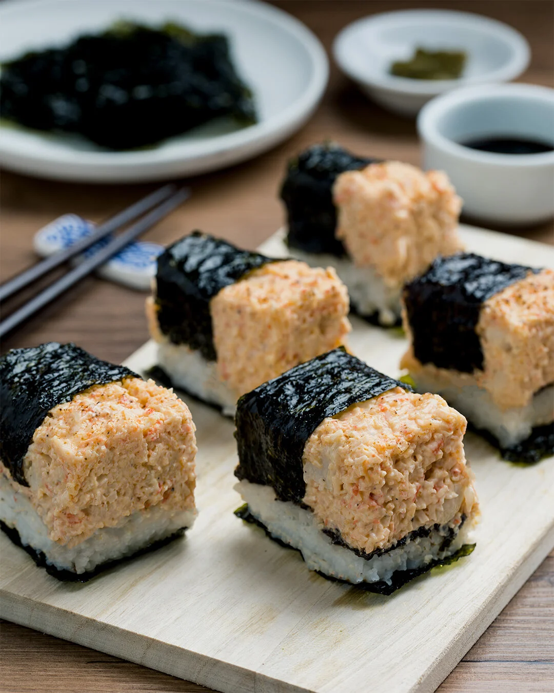 Six pieces of sushi topped with a seasoned mixture, wrapped with nori on a white wooden platter, with soy sauce, wasabi, and pickled ginger in the background.