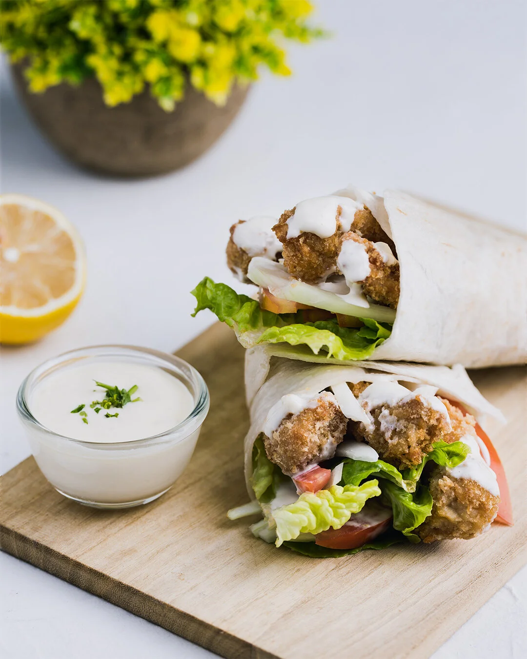 Two chicken wraps with lettuce, tomato, and creamy sauce on wooden board, served with a side of white dressing garnished with herbs, lemon half, and a plant in the background.
