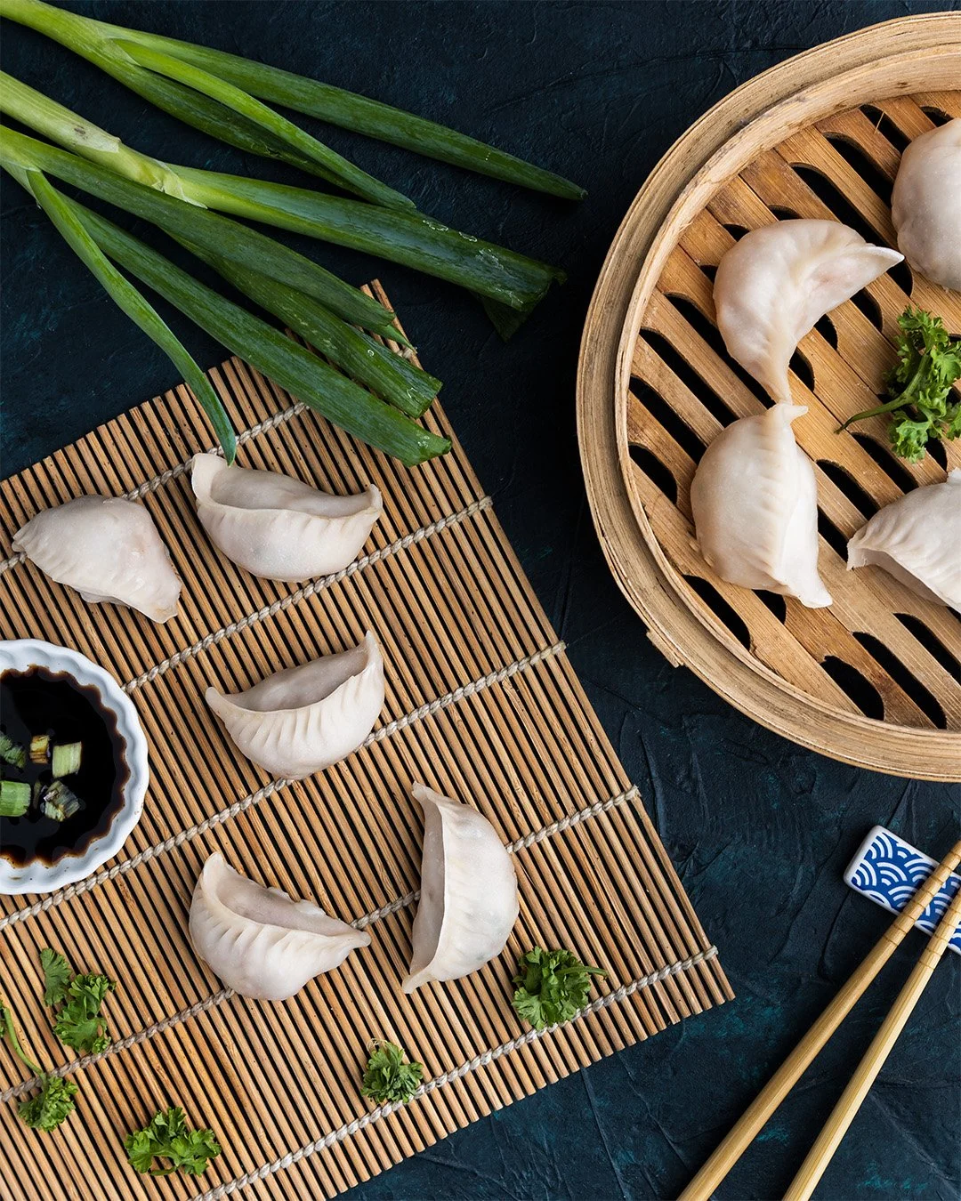 Steamed dumplings arranged on bamboo mats and a round bamboo steamer with additional dumplings and some green onions and cilantro, on a dark surface with chopsticks and soy sauce.
