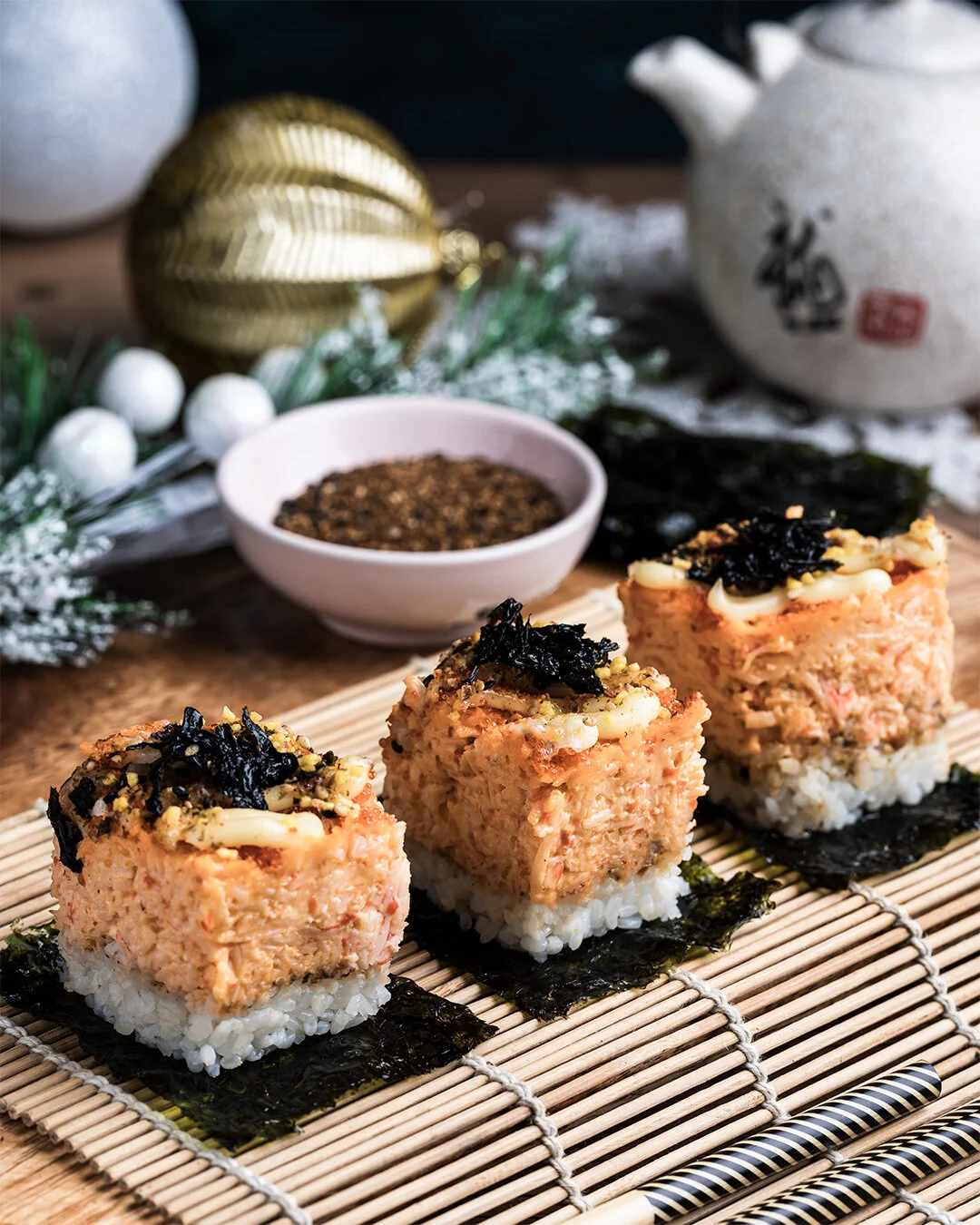 Three pieces of sushi with a topping of black seaweed and mayonnaise, placed on a wooden sushi mat with a background of Christmas decorations and dishware.