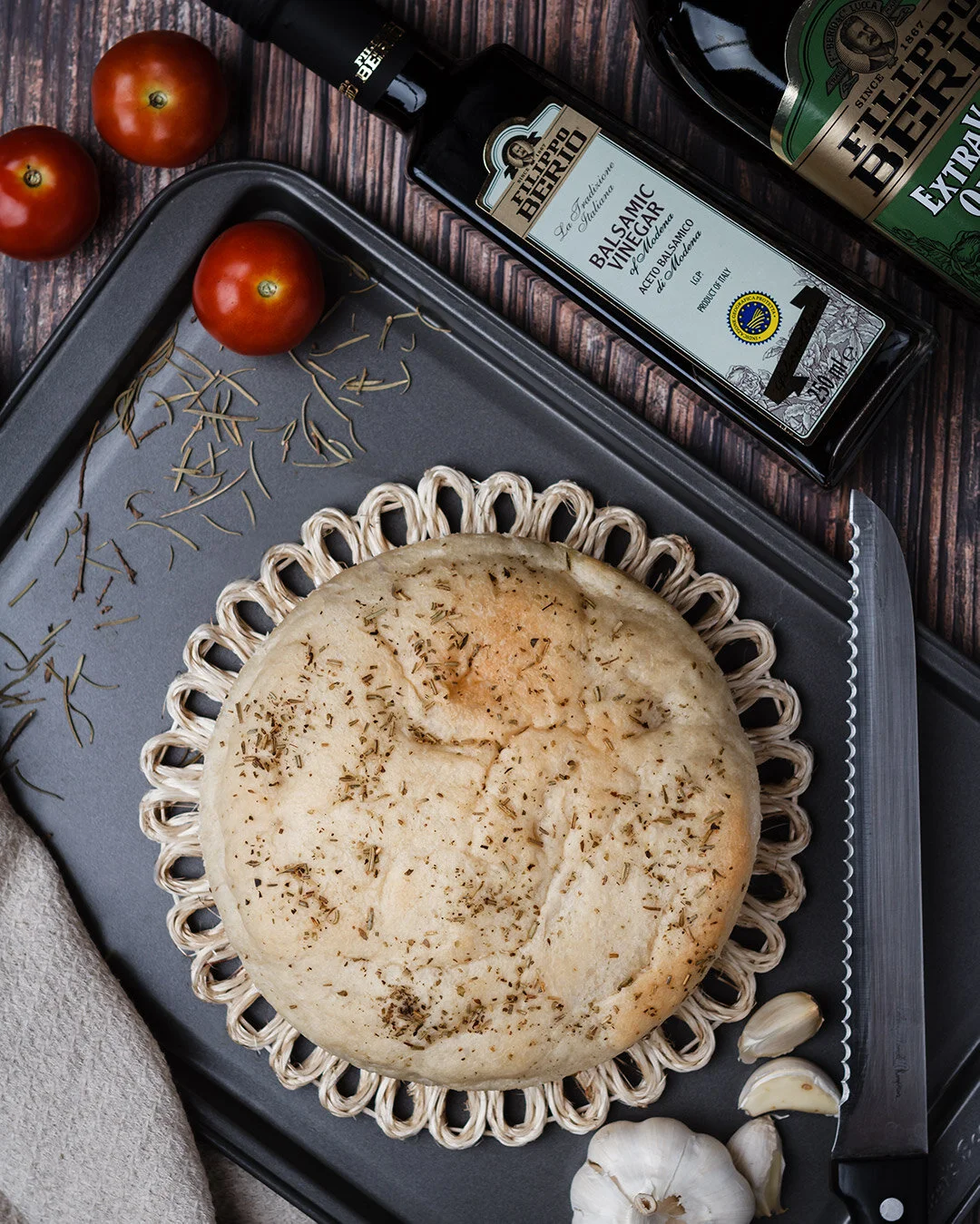 A round focaccia bread topped with herbs on a woven placemat. Surrounding it are tomatoes, garlic cloves, and sprigs of rosemary. There are bottles of balsamic vinegar and extra virgin olive oil nearby.