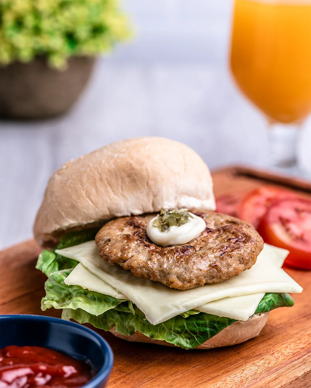A close-up of a burger with a beef patty, lettuce, cheese, a dollop of mayonnaise with pickles, and a sandwich bun on a wooden board, with sliced tomatoes, a small bowl of ketchup, and a glass of orange juice in the background.