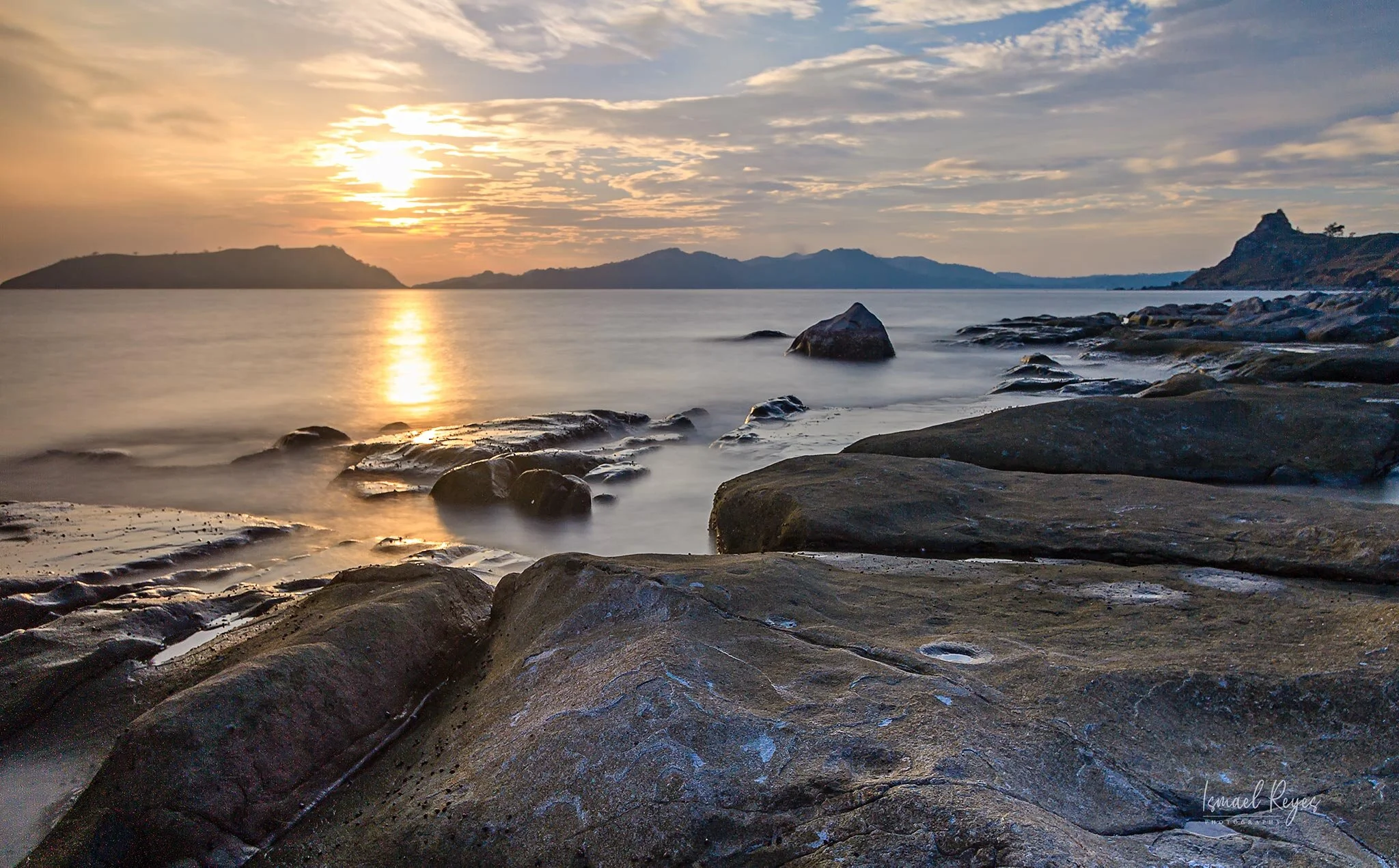 A serene sunset over a calm body of water with rocks along the shore and distant mountains in the background.