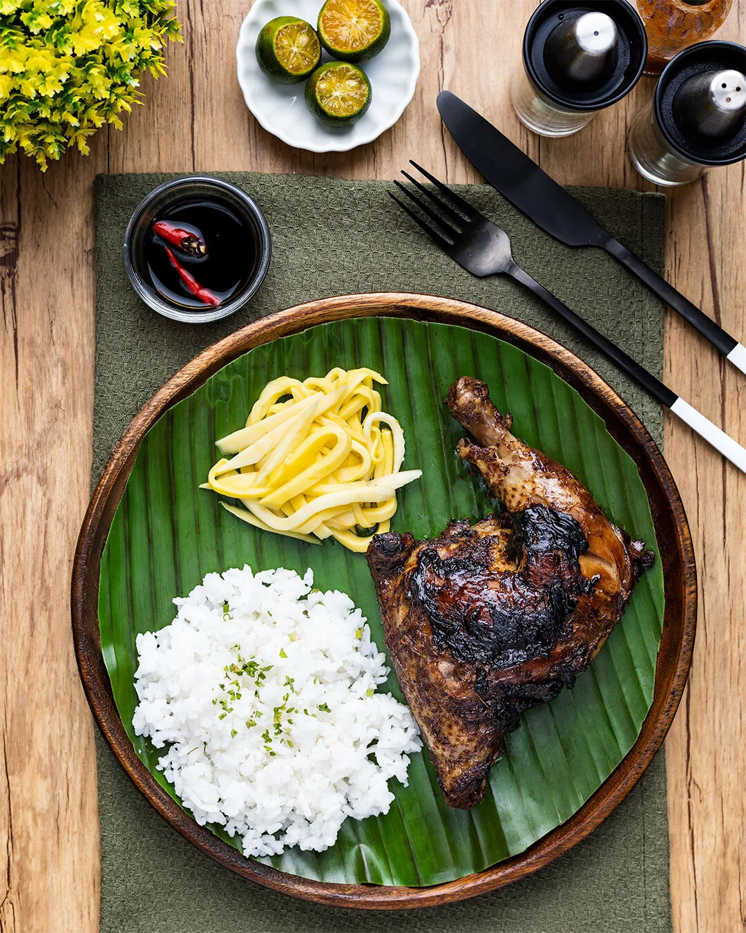 A plate featuring grilled chicken with dark char marks, served with white rice and yellow noodle salad on a green banana leaf atop a wooden table. Surrounding the plate are three lime halves on a white dish, black soy sauce with red chili peppers, a 