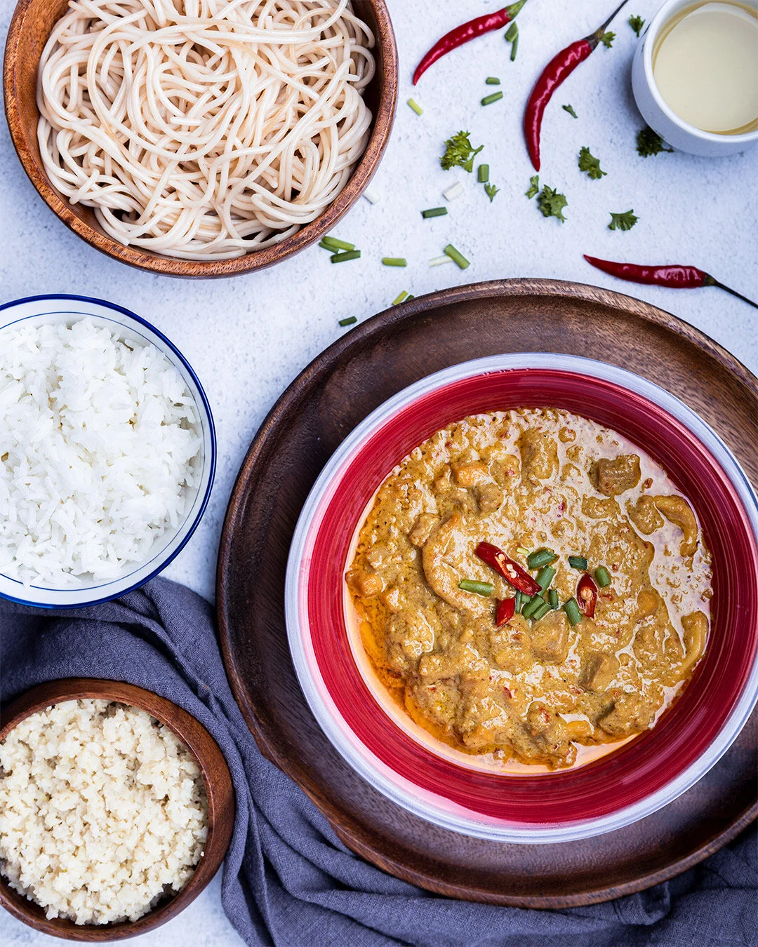 A plate of Thai green curry with chunks of meat, garnished with red chili slices and chopped green onions, served with bowls of cooked white rice, dried noodles, and crushed cashews on a white surface, with chili peppers and herbs nearby.