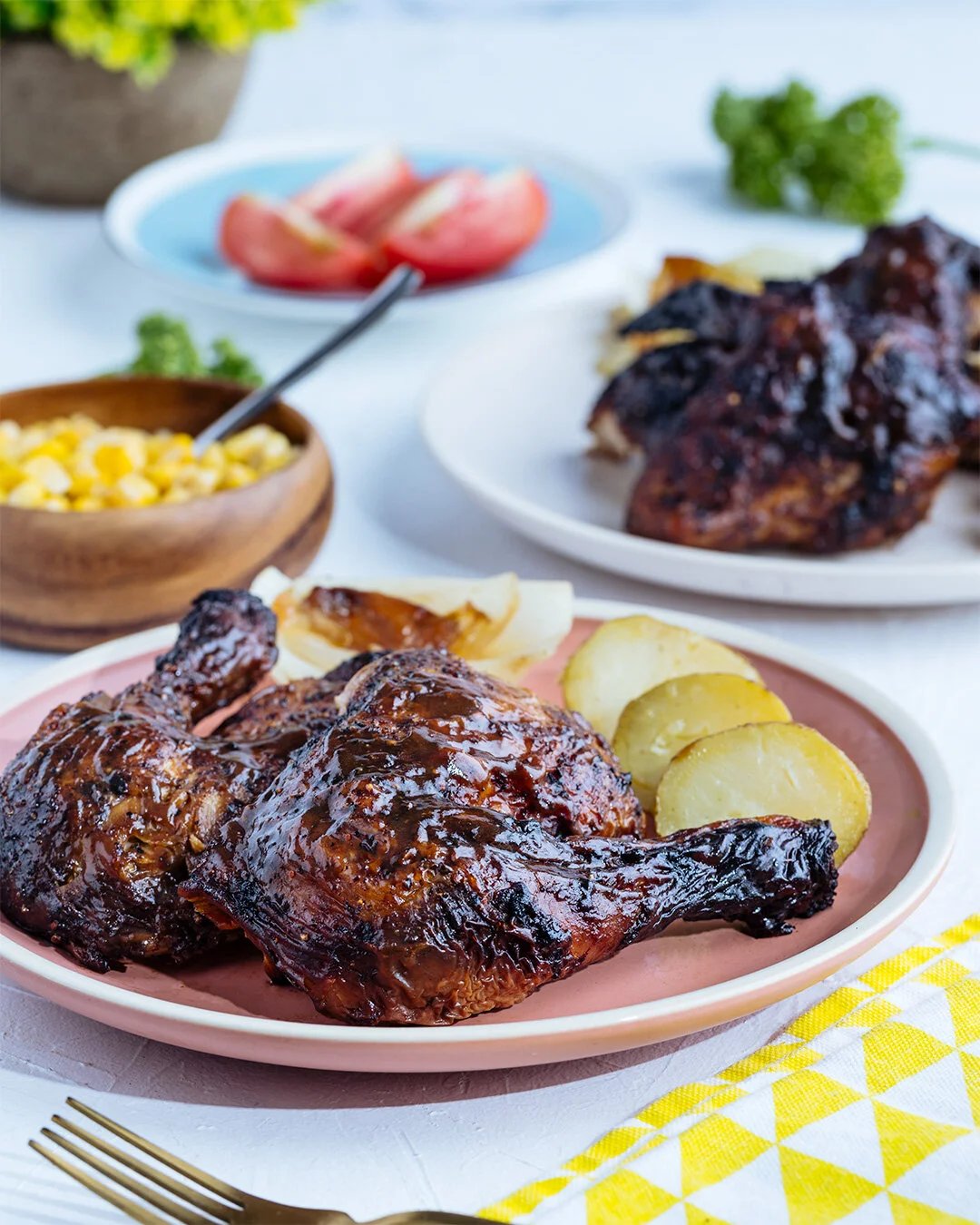 Close-up of a roasted chicken leg with potato slices on a pink plate, with additional roasted chicken in the background, a bowl of corn, and sliced tomatoes on a table.
