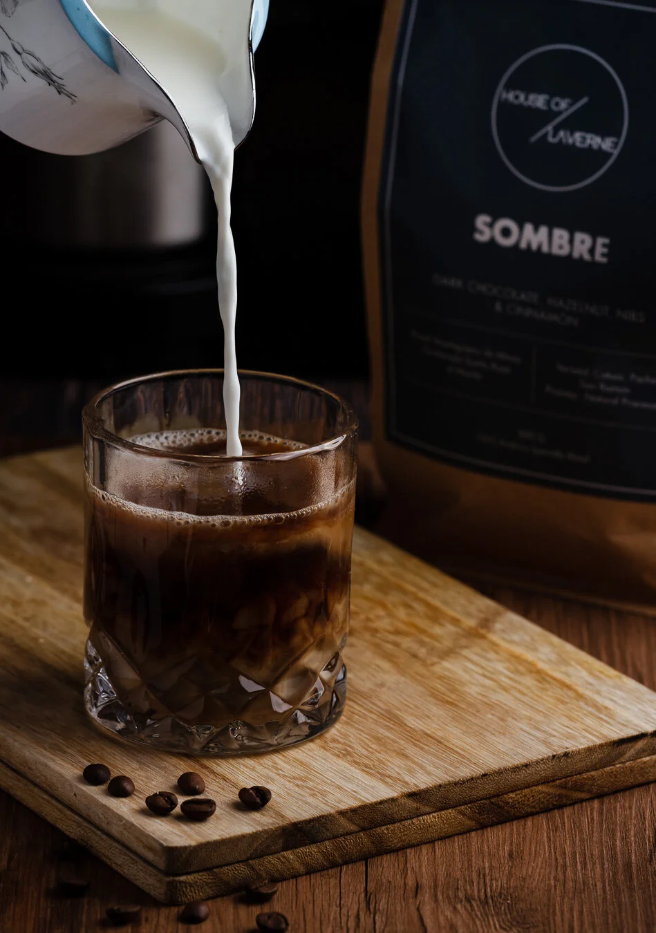 A glass of iced coffee on a wooden surface, with milk being poured into it from a metal pitcher, and coffee beans scattered around.