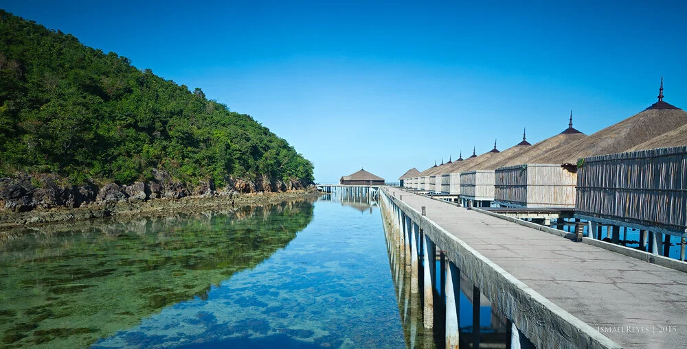 Overwater bungalows along a wooden pier extending over clear turquoise water, with a lush green hillside on the left and a bright blue sky overhead.