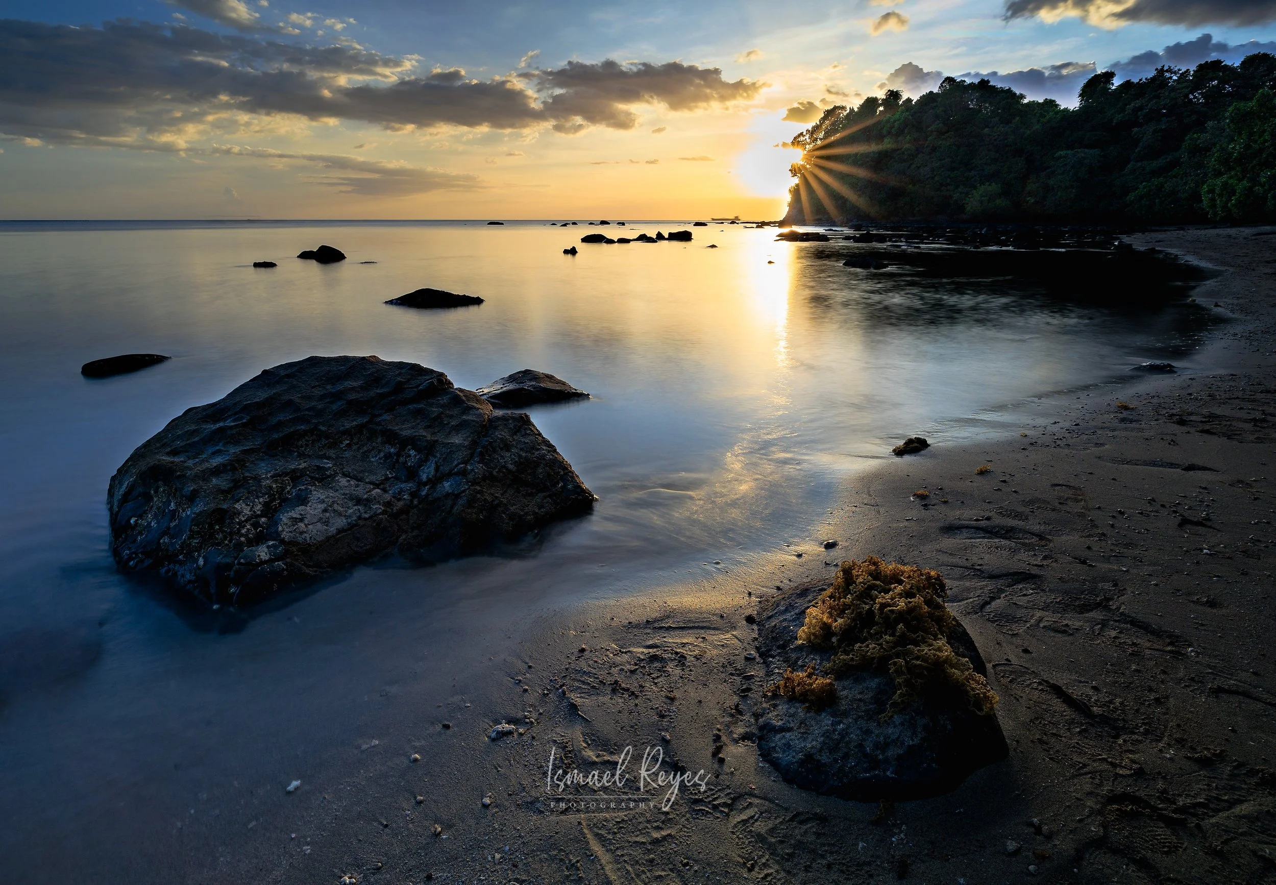 Sunset over a calm beach with rocks and small waves, and a tree-covered cliff on the right.