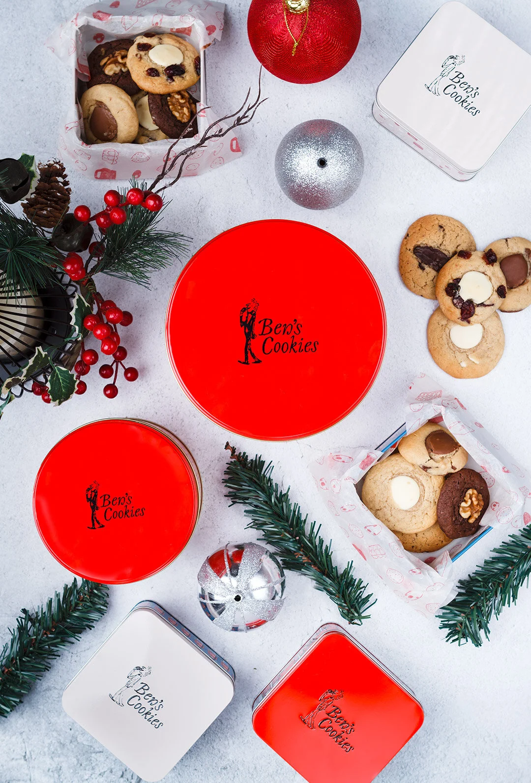 Holiday scene with decorated cookies, Christmas ornaments, and tins labeled 'Ben's Cookies' on a snowy surface.