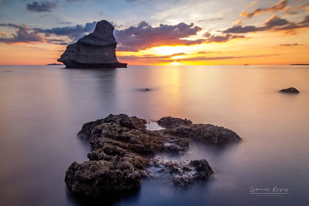 A sunset over the ocean with a large rock formation in the background and smaller rocks in the foreground. The sky has scattered clouds with colors of orange, purple, and yellow.