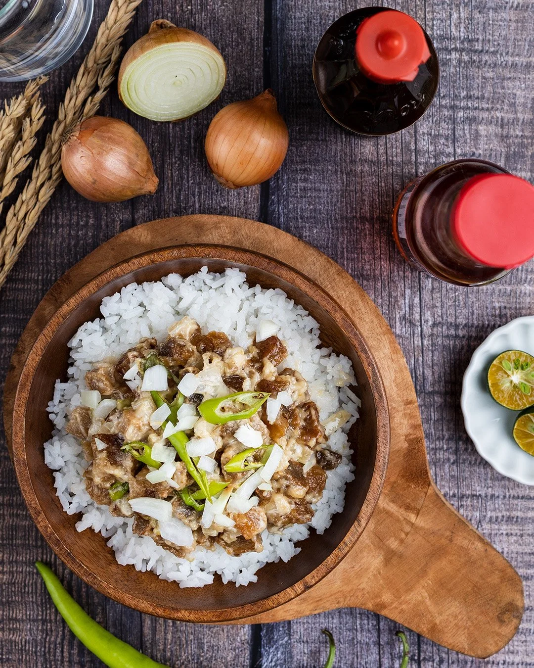 Cooked rice topped with a meat and vegetable mixture, garnished with chopped onions and green chili slices, served in a wooden bowl on a rustic wooden table with onions, soy sauce, vinegar, lime, and green chili nearby.