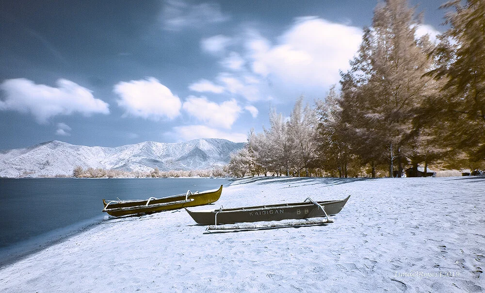 Snow-covered lakeside with two boats on the shore, trees, distant mountains, and partly cloudy sky.