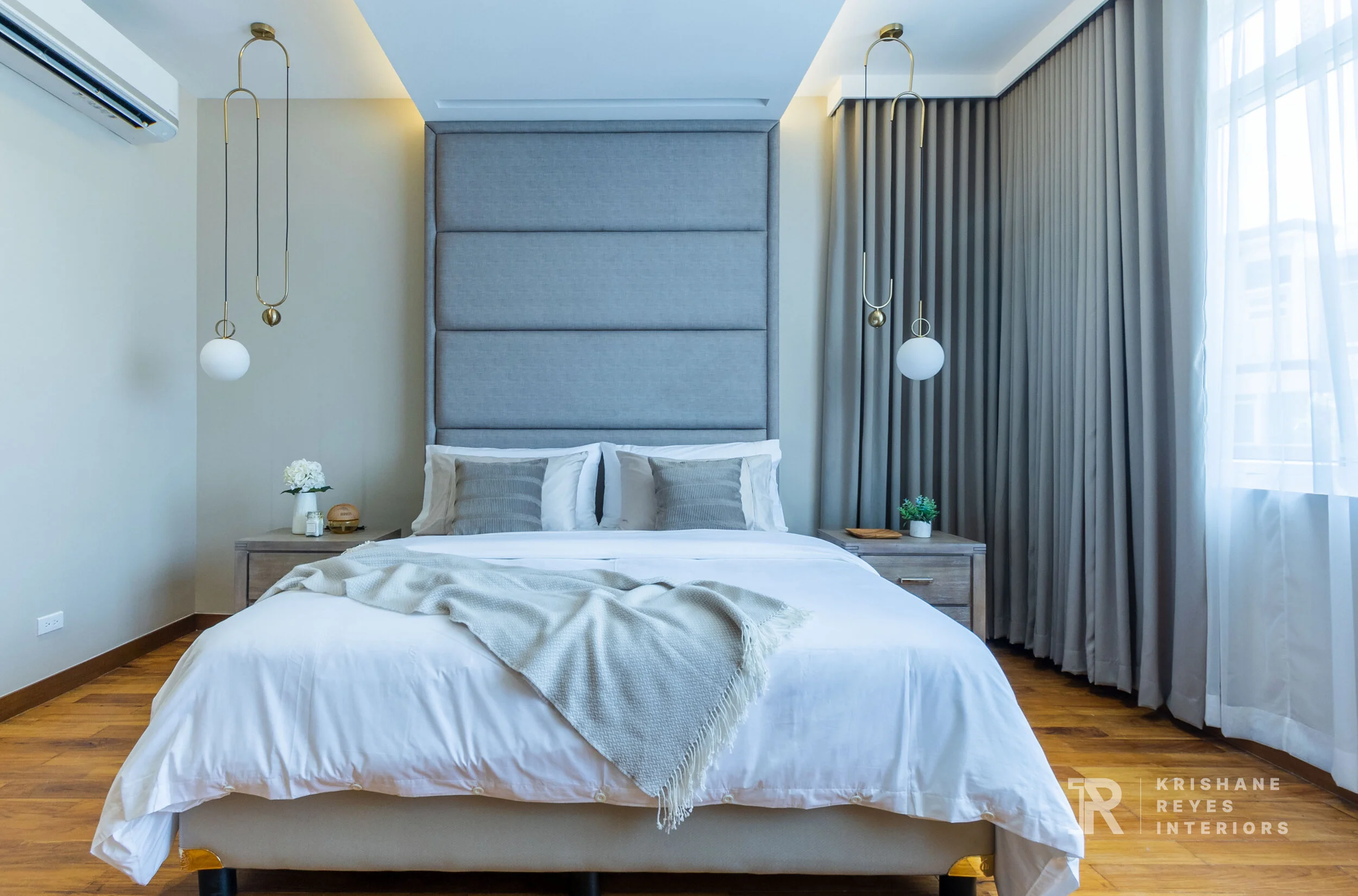 Modern bedroom with a large bed, gray upholstered headboard, white bedding, gray pillows, and beige throw blanket. Two nightstands with decor, hanging pendant lights, large curtains, window with sheer curtains, and wood flooring.