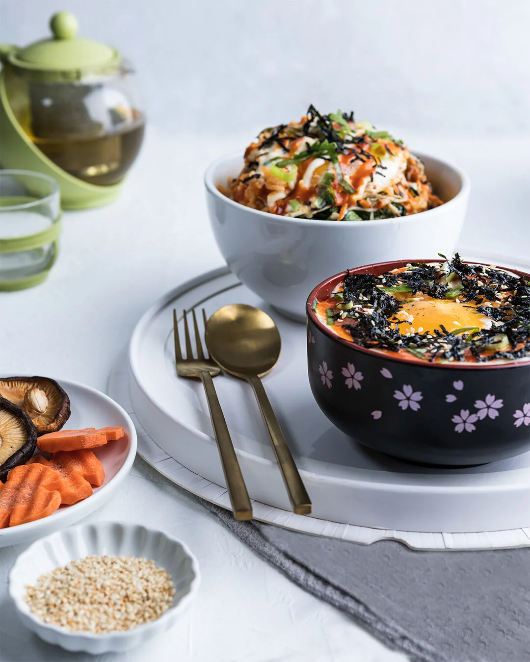 Bowl of ramen with an egg, seaweed, and green onions, placed on a white tray with a fork and spoon, surrounded by small bowls of vegetables and sesame seeds.