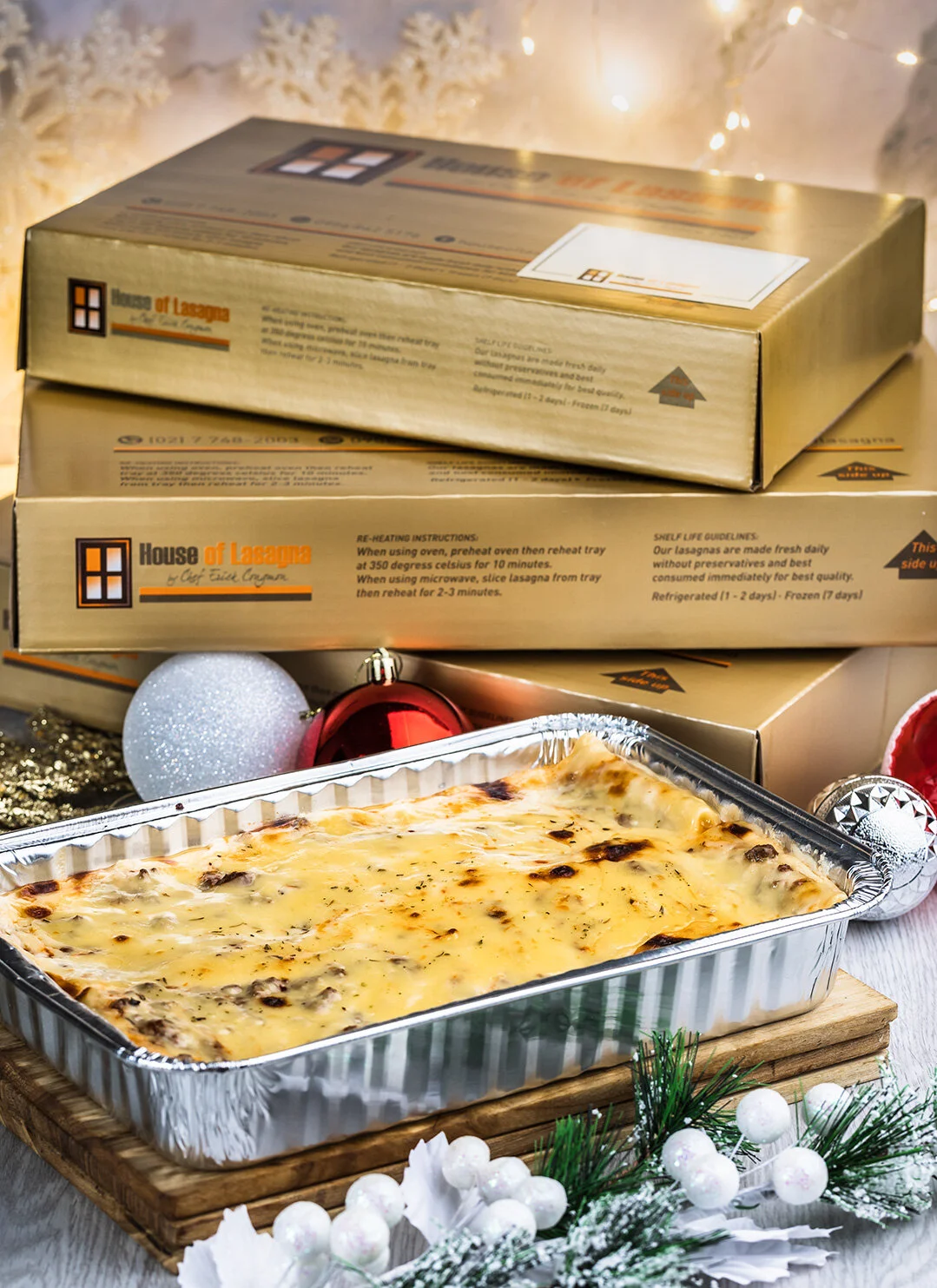 Baked lasagna in foil tray with holiday decorations and pizza boxes in the background.