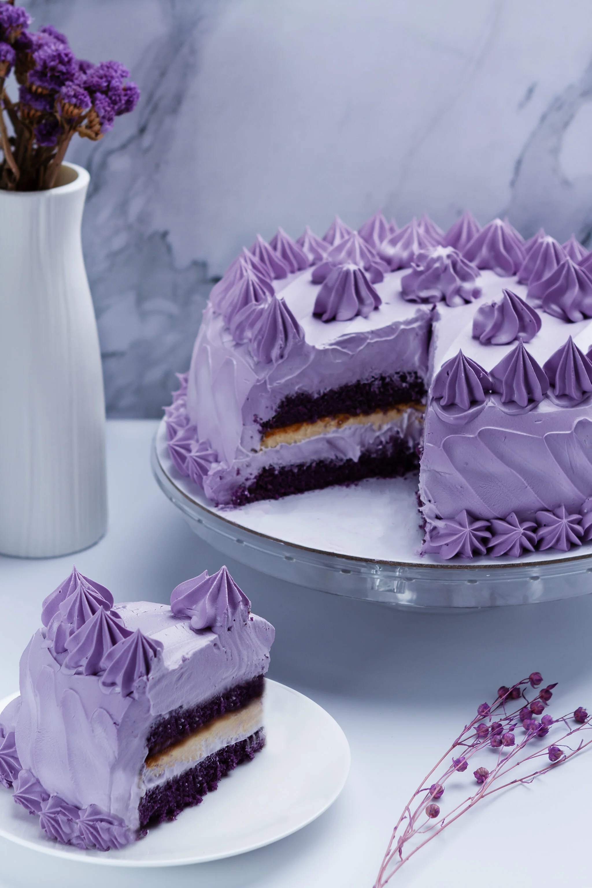 A purple layered cake with purple frosting and decorative piped frosting on top, partially sliced to reveal layers of purple cake, cream filling, and yellow cake, placed on a glass cake stand. A slice of the cake is on a white plate nearby, with a wh