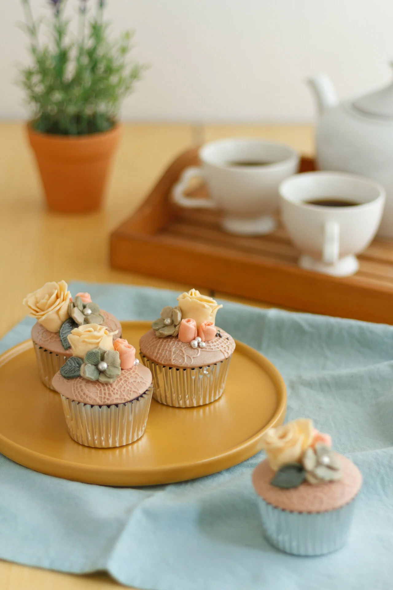 Decorative cupcakes with pastel floral frosting, placed on a yellow plate on a light blue cloth, with a tea set and a potted plant in the background.