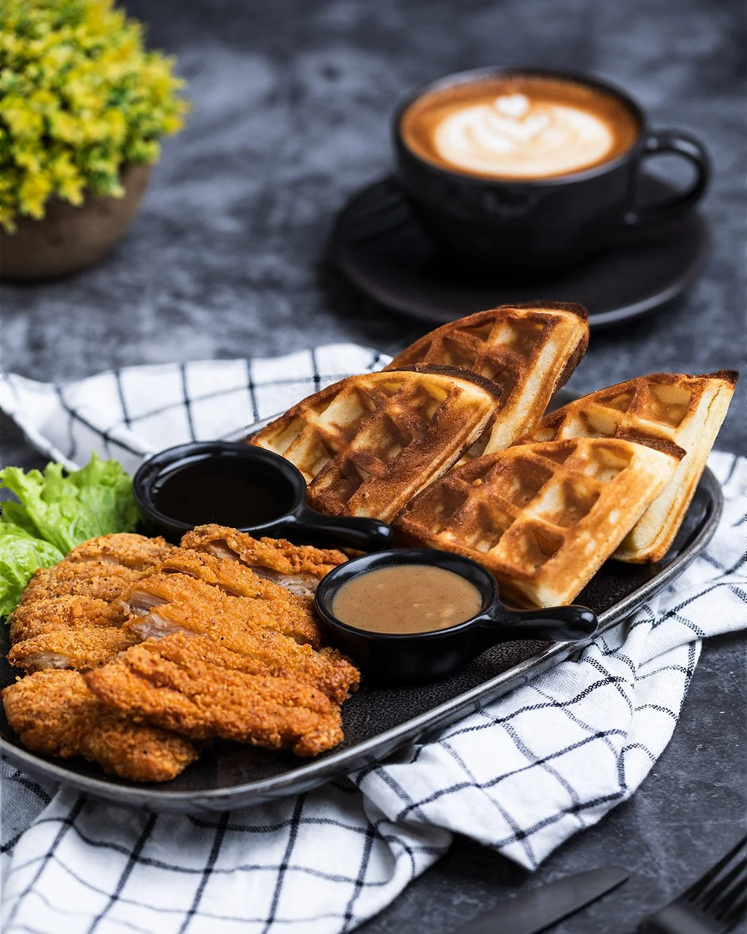 Fried chicken, waffles with syrup and sauces, lettuce, coffee in a cup, and a potted plant on a dark table.