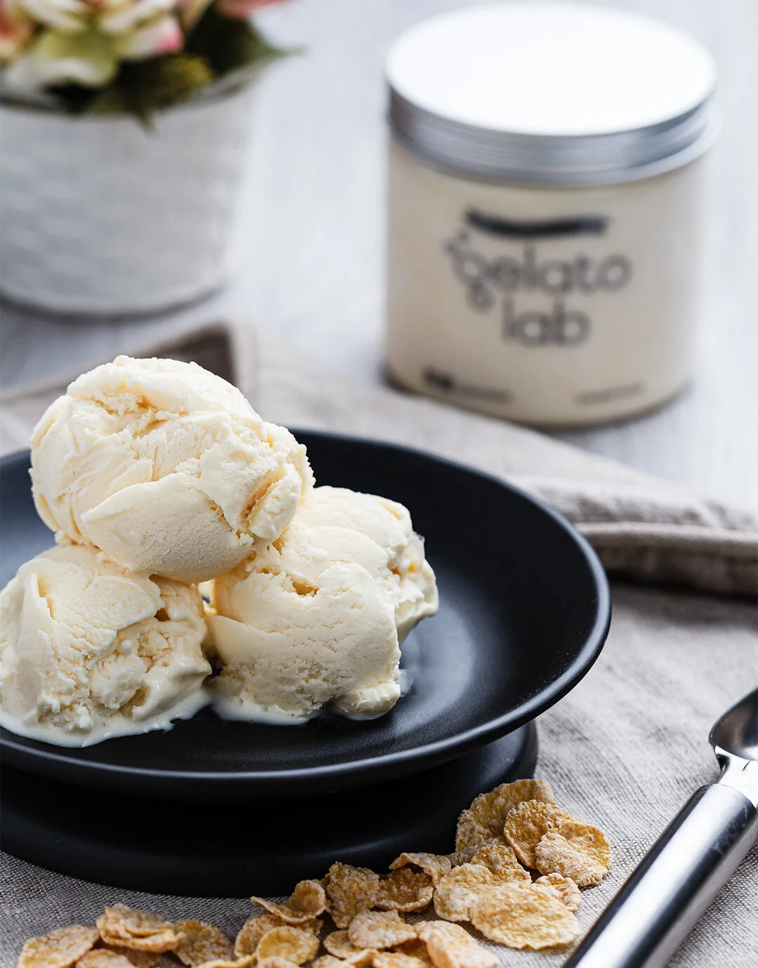Scoops of vanilla ice cream on a black plate, with cornflakes scattered nearby, on a textured beige cloth background.