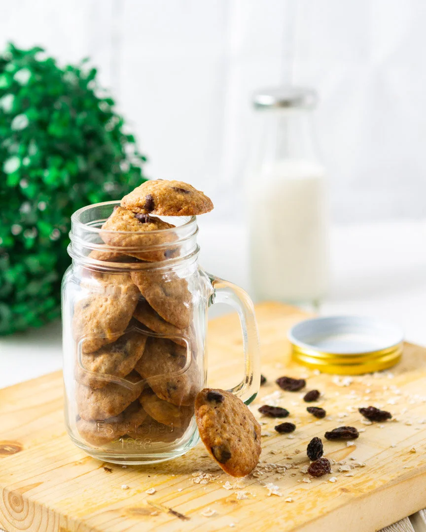 A glass jar filled with chocolate chip cookies on a wooden surface, with one cookie leaning against the jar, a few raisins and oatmeal scattered on the wood, a small open container of milk, and a bottle of milk in the background.
