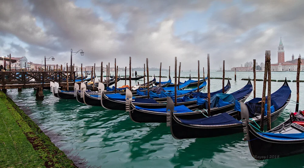 Gondolas docked in a canal in Venice, Italy, with a cloudy sky and historic buildings in the background.