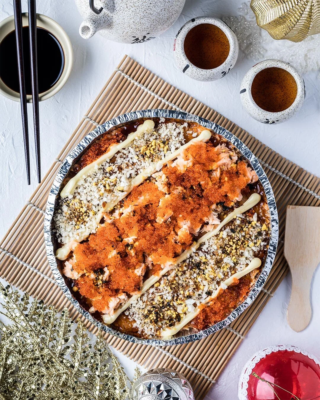 A festive holiday meal featuring a large sushi platter with various toppings, two cups of tea, a bowl of soy sauce, chopsticks, and holiday decorations like golden ribbons and a red ornament.