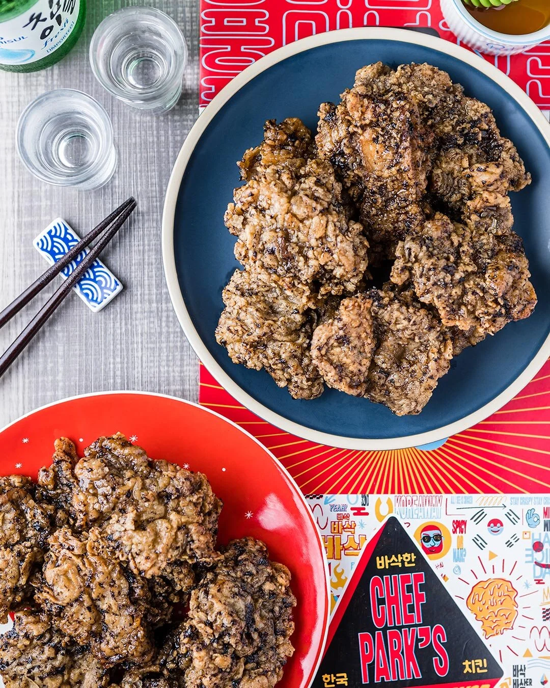Close-up of two plates of fried chicken on a table with glass cups, chopsticks, and a colorful paper placemat.