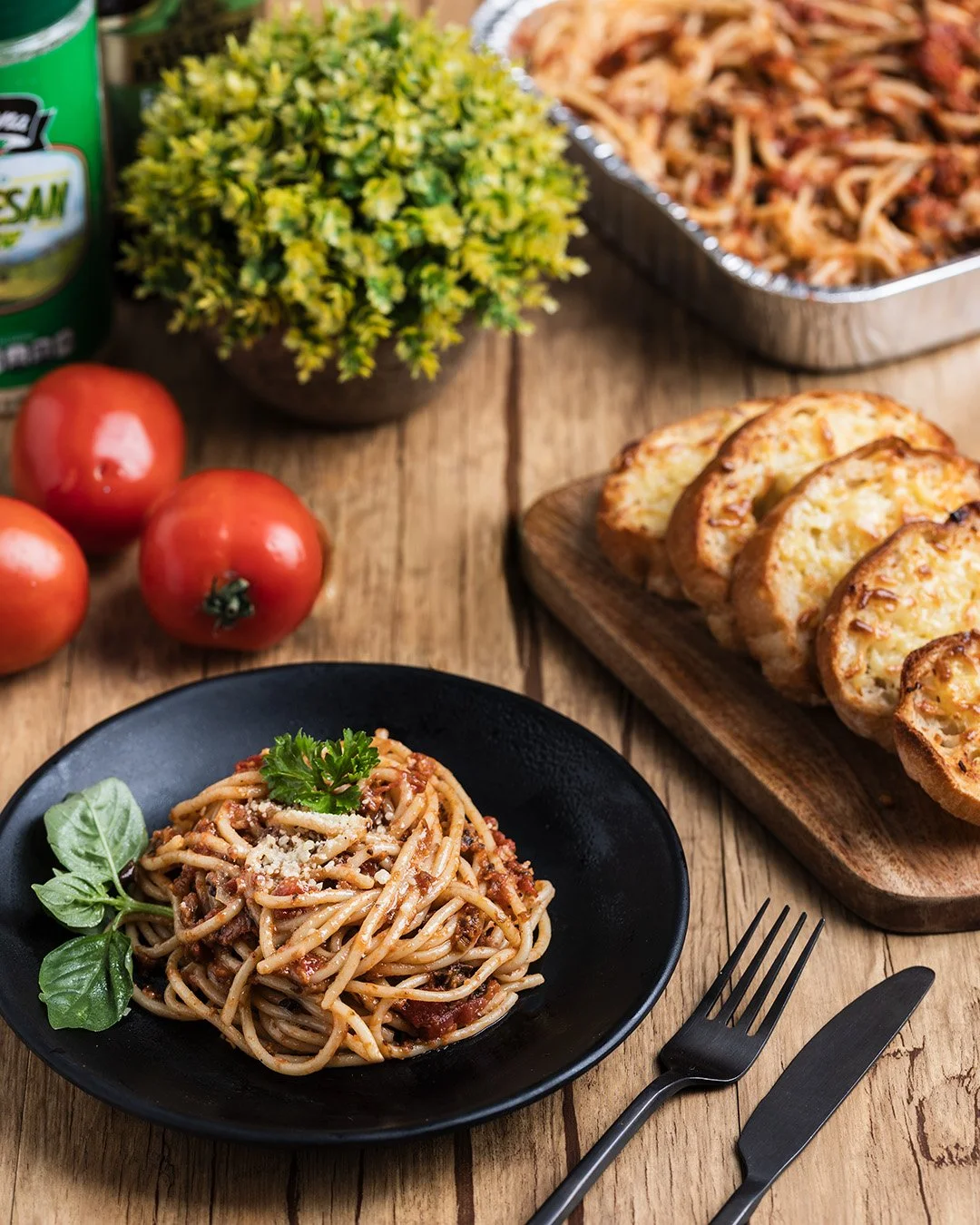 A plate of spaghetti with meat sauce garnished with parsley, alongside sliced garlic bread, tomato, canned vegetables, and a bowl of coleslaw on a rustic wooden table.