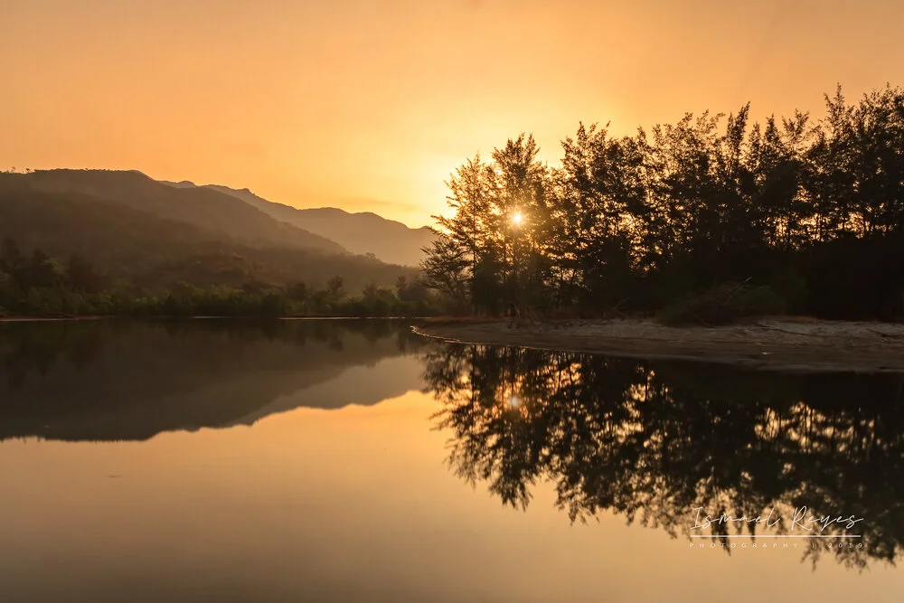Sunset over a calm river with trees and mountains reflected in the water.