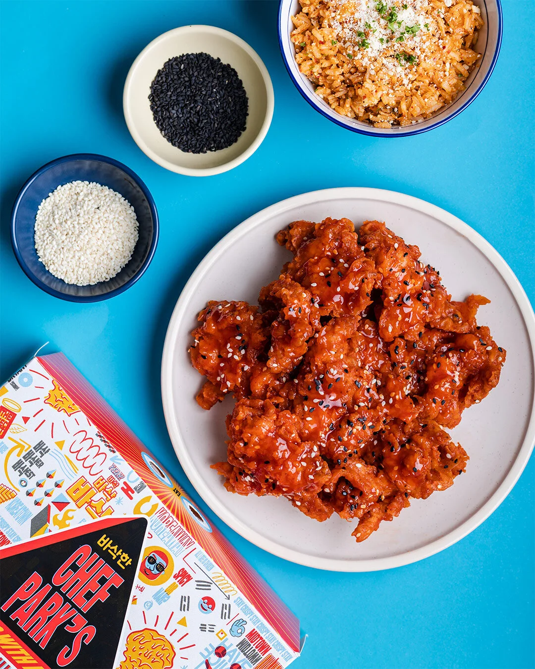 A plate of sweet and spicy Korean fried chicken topped with black and white sesame seeds, with side dishes of rice, black sesame seeds, and white sesame seeds on a blue background, along with a box of Chefs Park's Korean fried chicken.