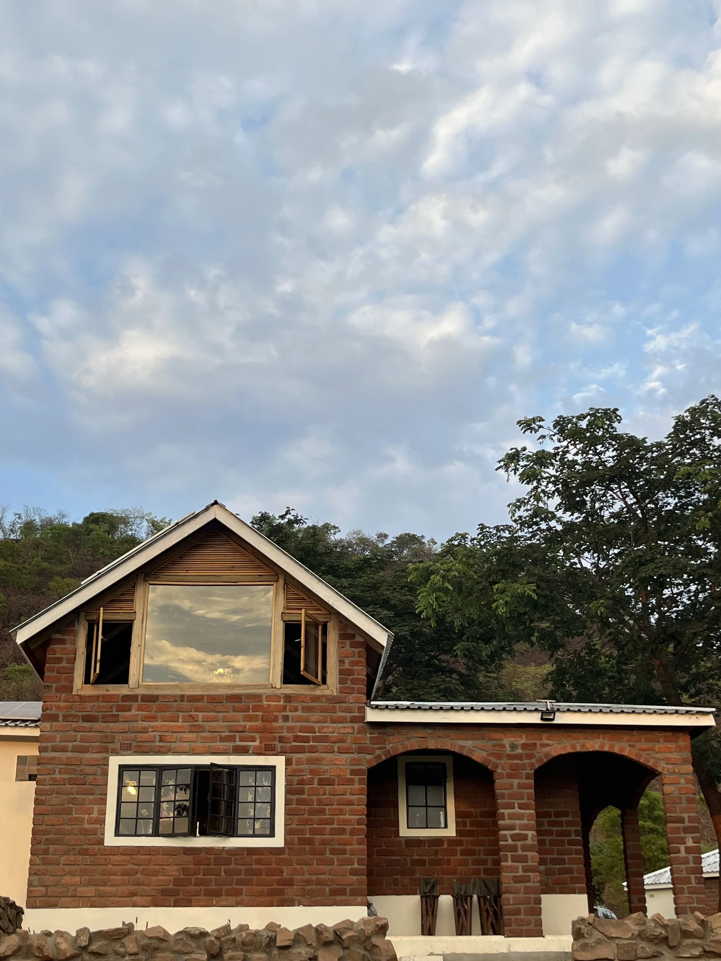 Image of Kampamba House's main building, showcasing its rustic charm with a thatched roof, natural stone walls, and wooden accents, nestled against the verdant backdrop of Leopard's Hill, indicating a serene getaway spot near Lusaka.