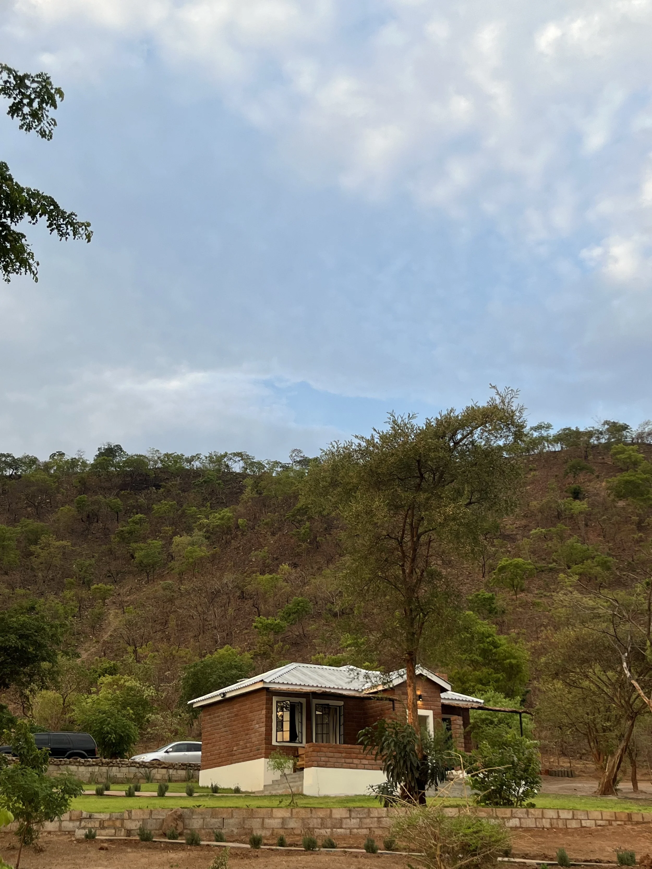 Image of a cozy, inviting chalet at Kampamba House, featuring a thatched roof, panoramic windows, and traditional decor elements, surrounded by lush greenery and offering a picturesque, tranquil retreat experience near Lusaka