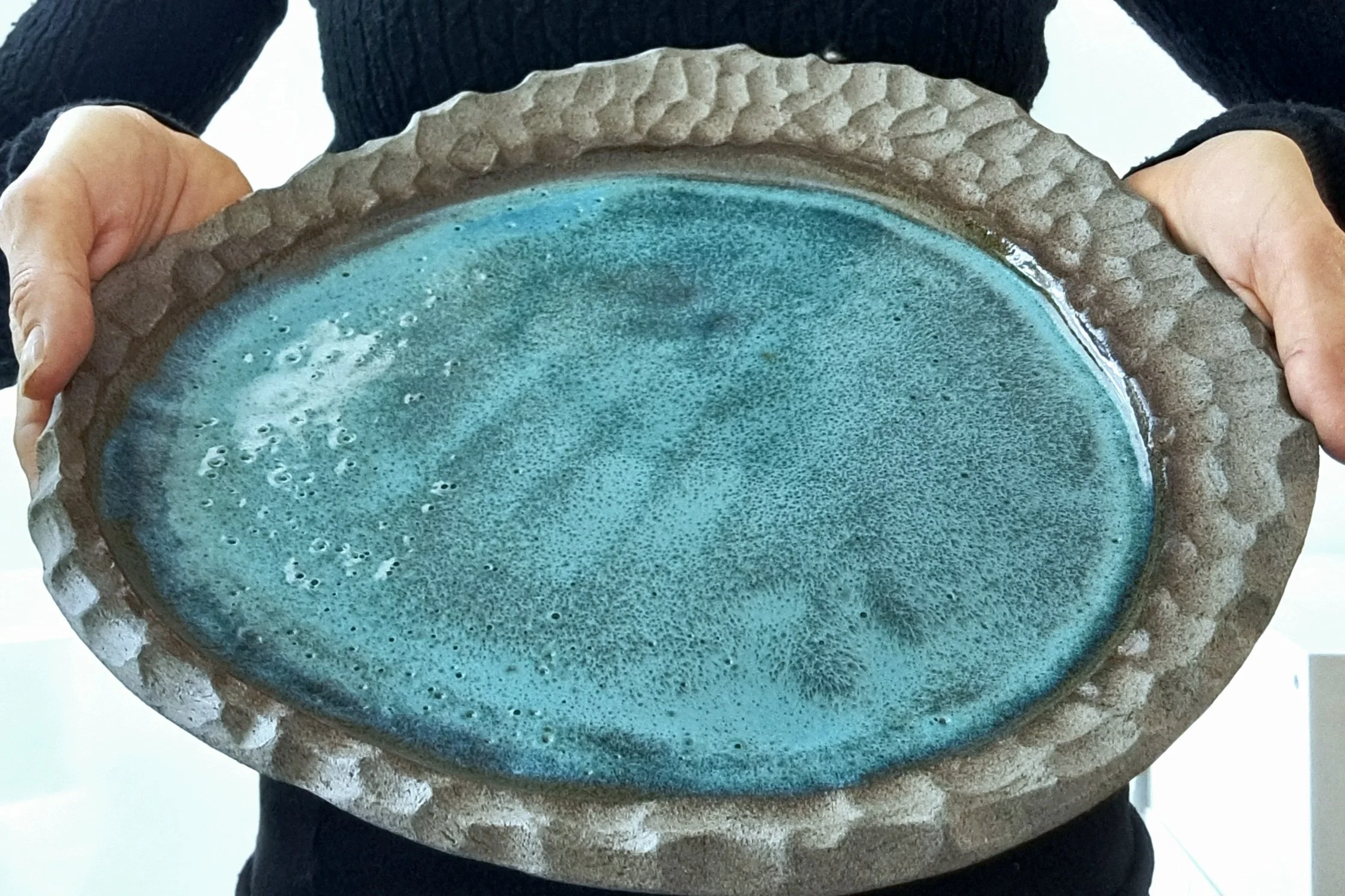 La persona tiene in mano una ciotola di ceramica con un interno di colore verde bluastro e superficie leggermente irregolare, indossa un maglione nero.