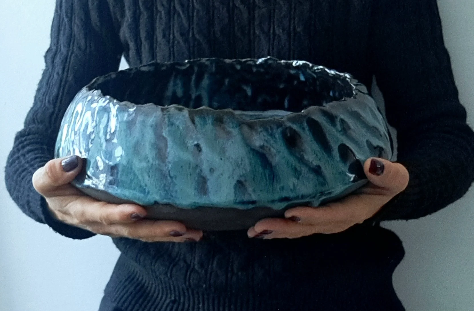 Persona che tiene in mano una ciotola grande blu con superficie irregolare e lucida, indossa un maglione nero a trecce.