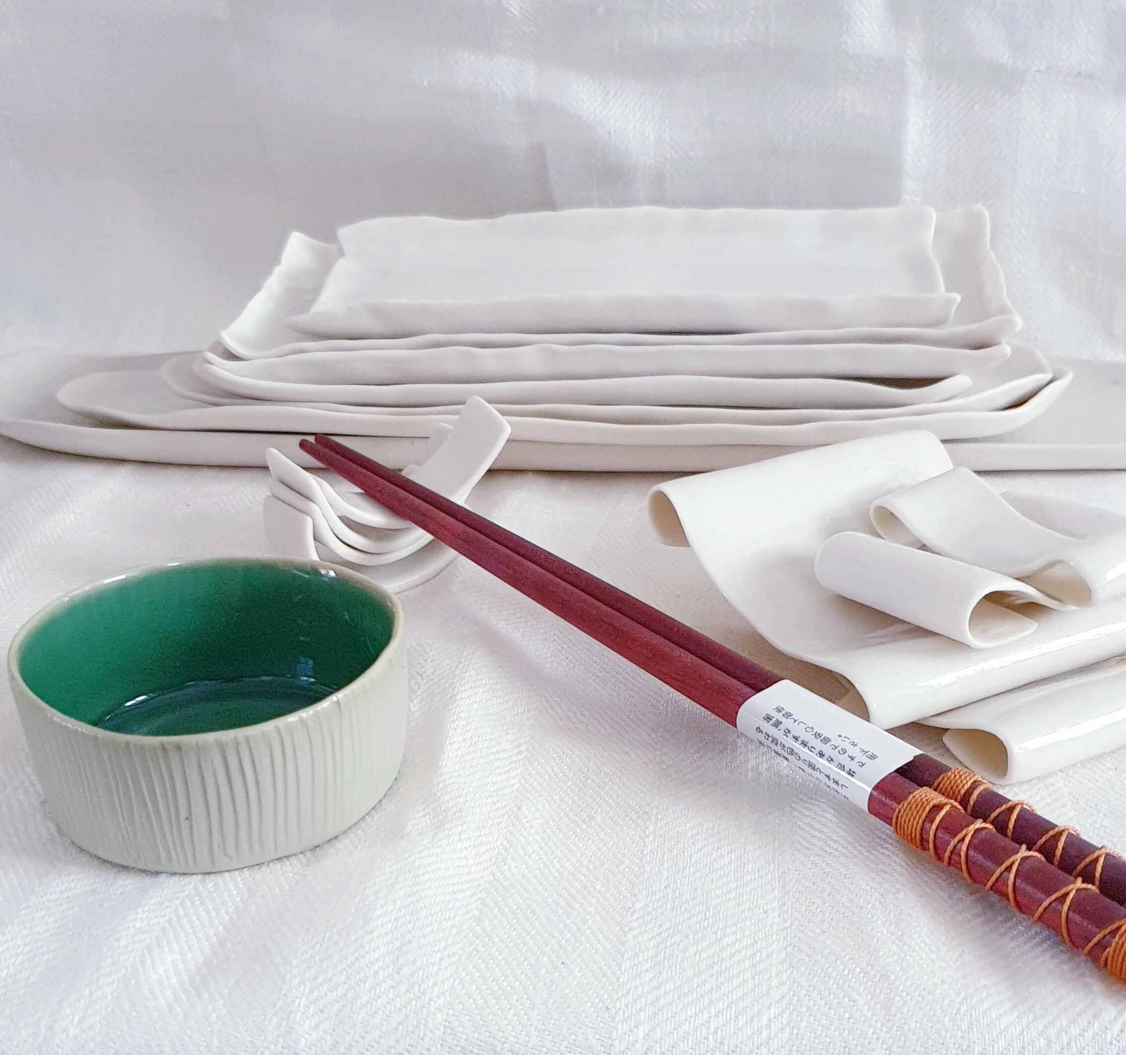 Set di ceramica per sushi con ciotola verde, bacchette rosse e portabacchette bianco. Piattini rettangolari impilati sullo sfondo.