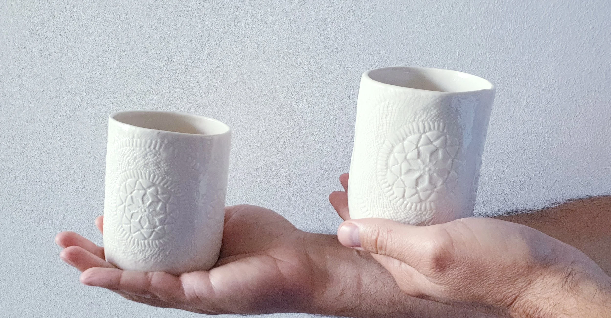 Due mani tengono due bicchieri di ceramica bianchi decorati con motivi geometrici, contro uno sfondo bianco.