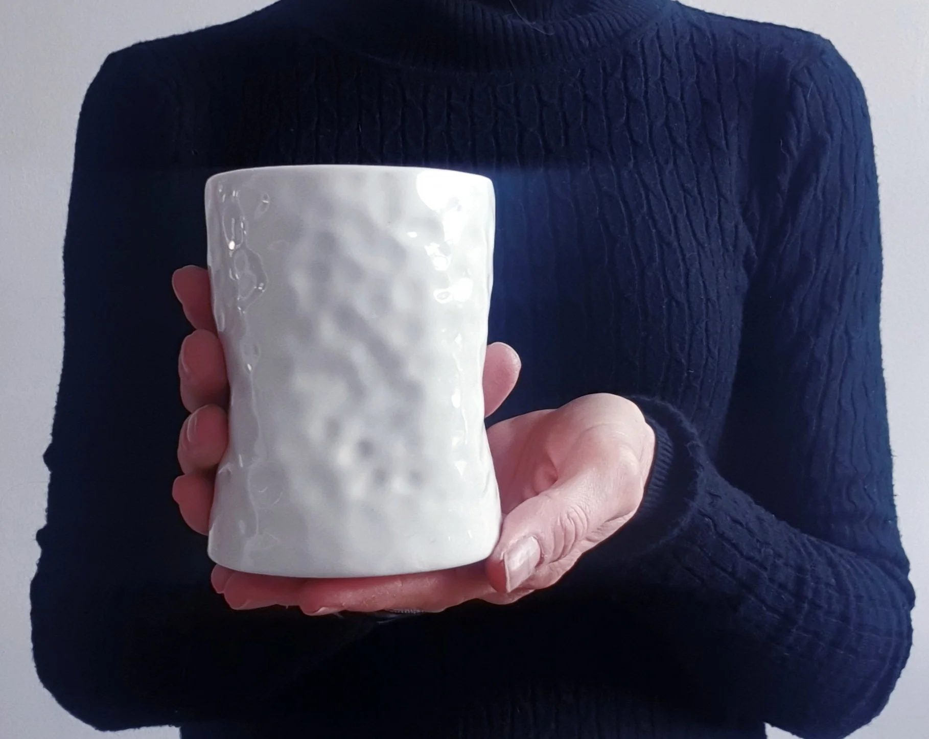 Persona che indossa un maglione blu scuro con cappuccio e tiene in mano una tazza bianca con superficie irregolare, visibile solo parte del volto e del corpo.