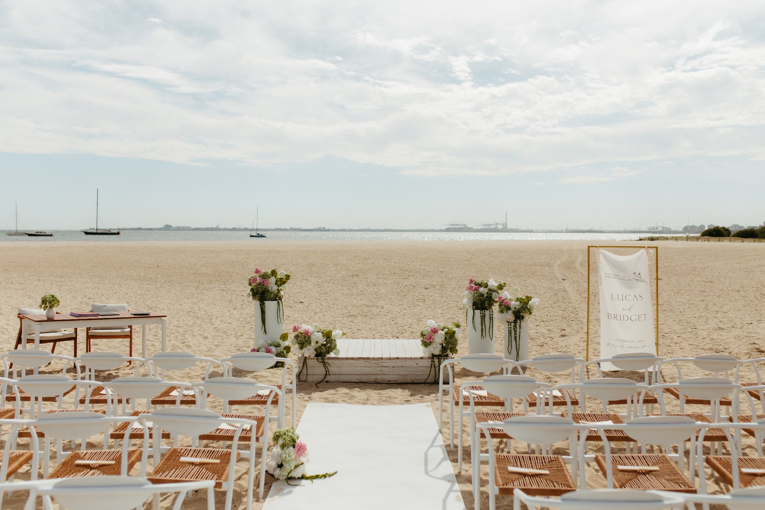 Outdoor wedding ceremony setup on a sandy beach with rows of white chairs, floral arrangements, an aisle, and a sign with names Lucas and Bridget, overlooking the ocean.